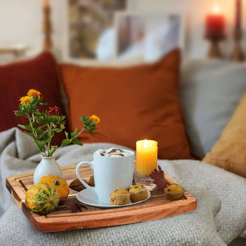 Acacia wood serving tray on a sofa with a blue mug, four muffins, two decorative pumpkins, and a vase of yellow flowers