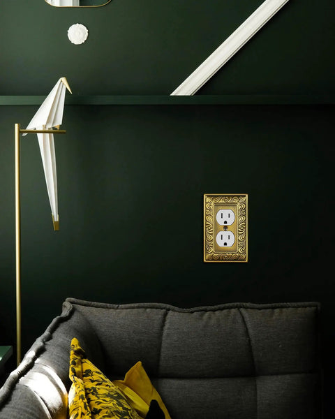 Six Pack Duplex Outlet Cover Plate With Paisley Pattern In Antique Brass Finish installed on a dark wall next to a sofa and lamp