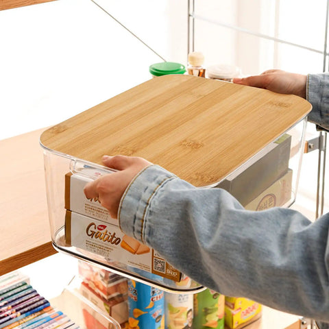 Person placing Clear PET Plastic Storage Bins With Removable Bamboo Lids And Grab Handles on a shelf, visible contents inside.