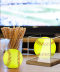 Clear Acrylic Softball Display Case With Wood Base For One Ball on table, next to glass with pretzels, with TV in background.