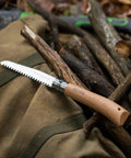 Badass Lightweight Folding Saw For Weekend Campouts And Garden Grind Cuts Branches Fast With Razor Sharp Carbon Steel Blade Secure Virobloc Lock And Natural Beechwood Handle on fabric with twigs in background.