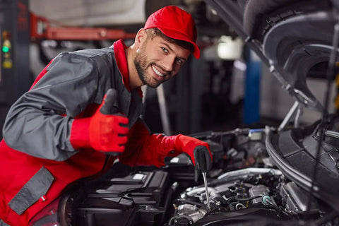 Mechanic in red uniform working under car hood in a garage, giving thumbs up.