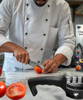Three Stage Kitchen Knife Sharpener For Steel And Ceramic Blades With Manual Whetstone And Polishing System Beginner Friendly Ergonomic Non Slip Base Get A Killer Razor Edge In Seconds beside chef cutting tomato on counter.