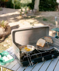 Portable stovetop camping toaster with four slices on a gas stove, stainless steel, ideal for outdoor cooking and picnics.