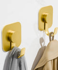 Heavy Duty Adhesive Wall Hooks In A Dope Four Pack, Stainless Steel Stick On Hangers With Badass Grip For Bathrooms And Kitchens, Waterproof And Super Fast To Put Up On Any Wall Or Door Without Drilling, Classic Gold Finish hanging towels on a white wall.