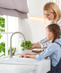 Mother and child washing vegetables under a kitchen faucet, with a plant on the windowsill.