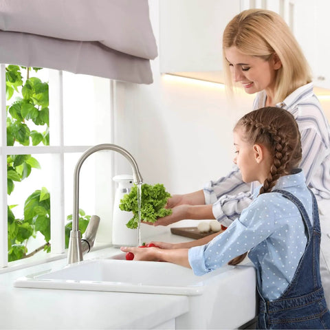 Mother and child washing vegetables under a kitchen faucet, with a plant on the windowsill.