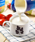 White ceramic mug with black leaf motif, milk being poured into it on a checkered cloth.