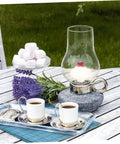 Oil lamp on outdoor table surrounded by flowers, marshmallows, and coffee cups, illustrating 10 Piece Cotton Wick Set For Oil Lamps Kerosene And Propane Lanterns Gas Lamps Outdoor Camping And Tiki Torches Round Design Portable And Easy To Replace Great For Picnics Road Trips And Backyard Gatherings.