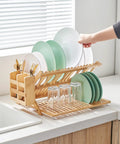 Heavy Duty Bamboo Dish Drying Rack Three Tier Collapsible Wooden Drainer With Utensil Holder holding plates and glasses on kitchen counter.