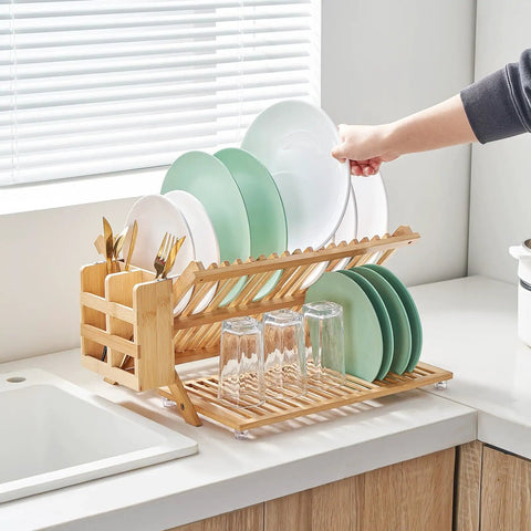 Heavy Duty Bamboo Dish Drying Rack Three Tier Collapsible Wooden Drainer With Utensil Holder holding plates and glasses on kitchen counter.