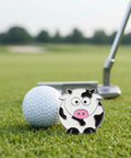Cow Shaped Golf Ball Marker With Hat Clip Cute Durable Metal Golf Accessory For Women Golfers Fun Gift Idea next to golf ball on green.