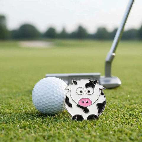 Cow Shaped Golf Ball Marker With Hat Clip Cute Durable Metal Golf Accessory For Women Golfers Fun Gift Idea next to golf ball on green.