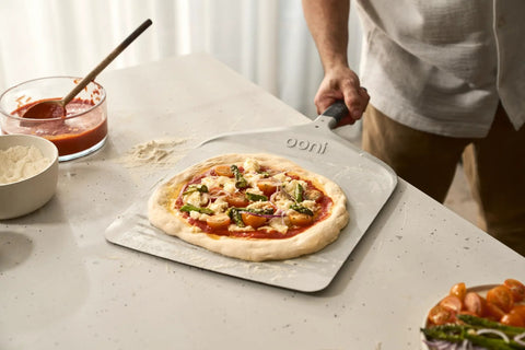 Front view of a 12-inch aluminum Ooni pizza peel with handle, pizza on the peel ready to bake.