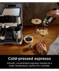 Person pouring cold-pressed espresso into glass beside coffee machine with beans on wooden table