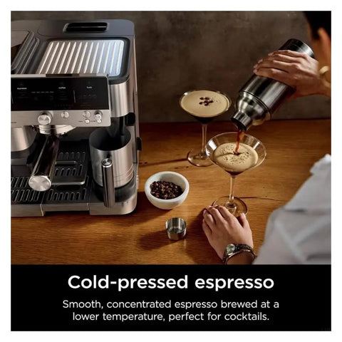 Person pouring cold-pressed espresso into glass beside coffee machine with beans on wooden table