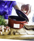 Person lifting lid of a red ceramic pot with sliced bacon and bread on a wooden board outdoors.