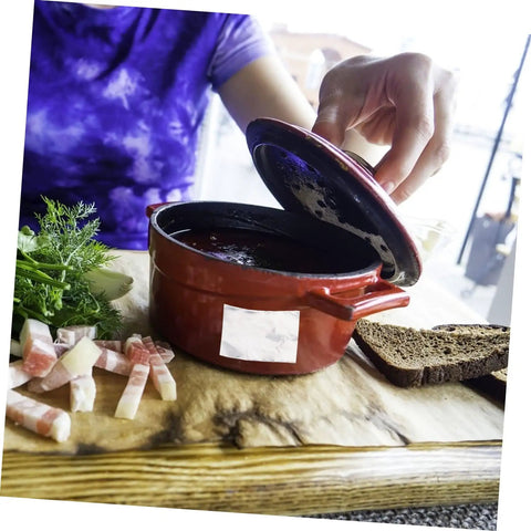 Person lifting lid of a red ceramic pot with sliced bacon and bread on a wooden board outdoors.
