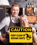 Two people smiling holding a Caution Keep Clear Of Moving Parts 8 By 12 Inch Aluminum Sign in a kitchen setting.