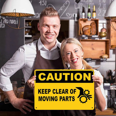 Two people smiling holding a Caution Keep Clear Of Moving Parts 8 By 12 Inch Aluminum Sign in a kitchen setting.