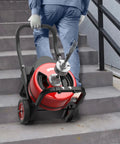 Heavy Duty 100Ft Electric Drain Auger With 750W Auto Feed Steel Core Cable being carried up stairs by a user for pipe cleaning.