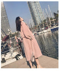 Woman in pink coat and beige shoes looking up at buildings near the water with boats and palm trees in the background.