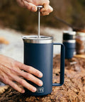 Person using Rambler 34 Oz French Press Coffee Maker With Grounds Control Filter Navy Double Wall Vacuum Insulation Keeps Your Brew Hot On The Go Easy Pour Durable Build For Everyday Boss Coffee Experience outdoors on wooden surface