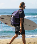 Man carrying a surfboard on the beach wearing 35L Waterproof Swim Backpack With Separate Shoes Compartment, ultra spacious purple sports rucksack