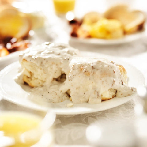Close-up of biscuits topped with creamy sausage gravy on a white plate, with breakfast items blurred in the background.