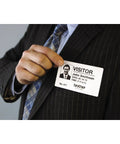 Man in a suit holding a visitor badge, showcasing Ultra Fast Network Label Printer With Bluetooth And Ethernet Wireless Connectivity Slick Monochrome Printing Up To One Hundred Ten Labels Per Minute Dead Simple Setup For Professional Workflows.