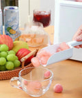No Fuss Countertop Ice Maker drops nine chill cubes into a clear glass, surrounded by fruit and drinks on a wooden table.
