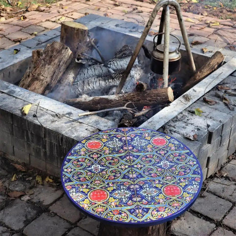Intricate round table with colorful mosaic pattern near a brick fire pit with kettle, logs, and smoke in outdoor setting.