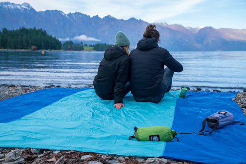 Couple sitting on Oversized Sandproof Beach Blanket 10x9 For 2 To 8 Adults by lakeside, blue and teal mat against mountain backdrop.