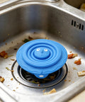 Drain Unclogger Flexible Bath Drain Cover Sealing Stopper in blue silicone resting in a kitchen sink with food debris.