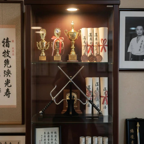 Dope Dual Blade Display Stand For Sai And Kama showcasing sai weapons in a display case with trophies and certificates.