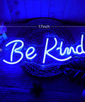 Blue 'Be Kind' neon sign, 17x7 inches, glowing on a textured background with leaves visible, neon wall decor with bright blue light