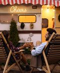 Couple toasting in front of camper, "Cheers" neon sign above, wood chairs, evening setting