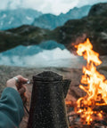 Person holding 3 Qt Coffee Boiler in Speckled Black by campfire, ideal for quick brews and rapid heat absorption, 12-cup capacity