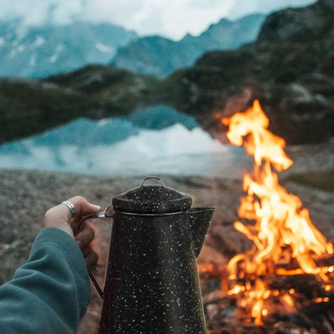 Person holding 3 Qt Coffee Boiler in Speckled Black by campfire, ideal for quick brews and rapid heat absorption, 12-cup capacity