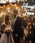 Bride and groom under 'Always & Forever' neon sign at wedding ceremony with floral decorations and soft lighting.