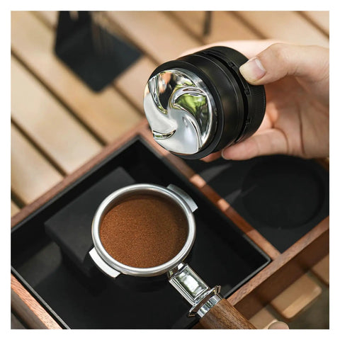 Pro Level Espresso Coffee Distributor And Tamper Set 58.35mm in hand above a portafilter with ground coffee on a wooden surface.