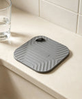 Shower Drain Cover Tub Stopper Square Silicone Floor Drain Cover in gray, placed on a countertop beside a glass of water.