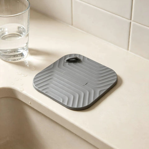 Shower Drain Cover Tub Stopper Square Silicone Floor Drain Cover in gray, placed on a countertop beside a glass of water.