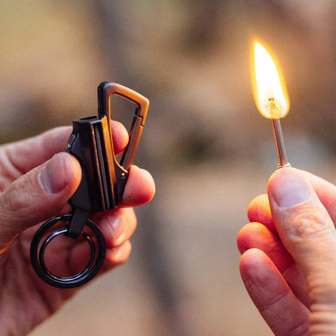 Hand holding Permanent Match Bottle Opener Keychain, Reusable Survival Fire Starter Striker Lighter, lit flame, charcoal finish.