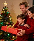 Family exchanging a gift in front of a Christmas tree, decorated with colorful lights and ornaments during a festive holiday gathering.