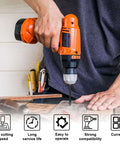 Front view of an orange cordless drill with bit installed drilling into a wooden board on a workbench; tools in background.