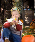 A child in royal costume excitedly holds Halloween candy outdoors, surrounded by pumpkins and spooky decor.