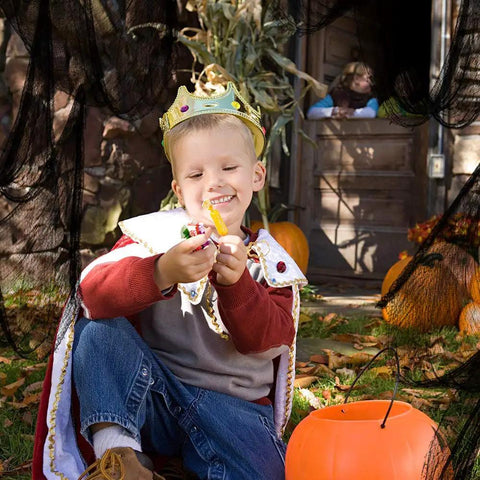 A child in royal costume excitedly holds Halloween candy outdoors, surrounded by pumpkins and spooky decor.