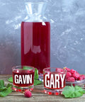 Clear glasses with names Gavin and Gary, filled with red juice next to a full carafe and fresh raspberries on a wooden table.