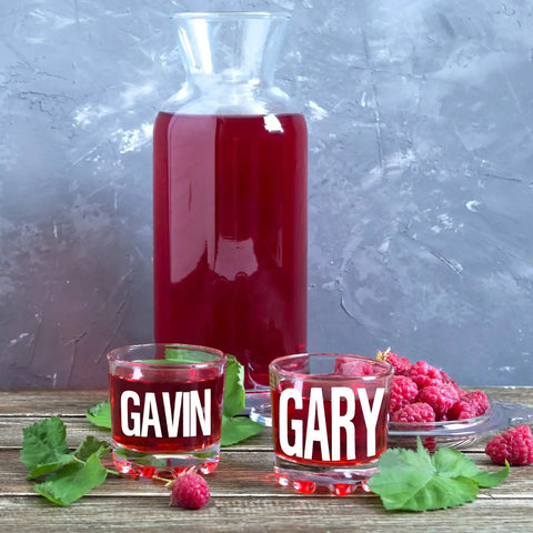 Clear glasses with names Gavin and Gary, filled with red juice next to a full carafe and fresh raspberries on a wooden table.